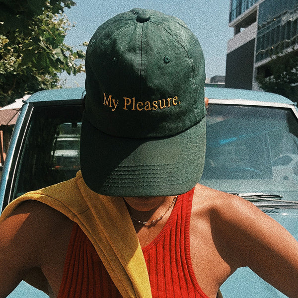 forest green baseball cap with 'my pleasure' written in yellow serif type by Yokoko. The hat is worn by a female model in a red tank top in front of a blue car.